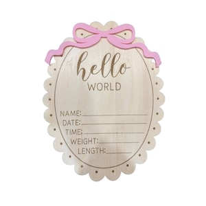 May include: An oval wooden birth announcement sign with a scalloped edge and a pink bow at the top. The sign features the text hello WORLD and blank lines for NAME, DATE, TIME, WEIGHT, and LENGTH. The wood has a light natural grain finish.