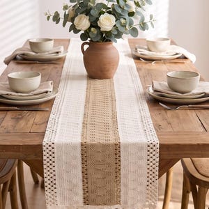 May include: A long rectangular table runner featuring a central tan woven panel flanked by cream-colored lace sections with intricate patterns. The runner ends in a row of long, cream-colored fringe tassels hanging off the edge of the wooden table.