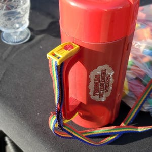 May include: A red plastic thermos with a handle and a multi-colored striped carrying strap. The lid features a circular sticker with an image of Thomas the Tank Engine and the text "Thomas says have a great day!" and a side label.