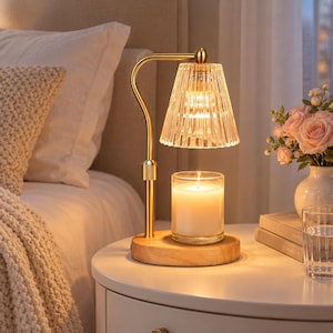 May include: A candle warmer lamp featuring a gold-toned metal arm, a round light-colored wooden base, and a clear, vertically ribbed glass shade. A glass jar candle with a lit wick sits on the base, illuminated by the warm glow from above.