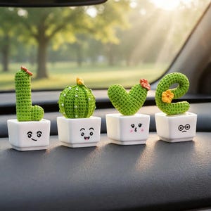 May include: Four green crocheted cactus figures in white square pots, each with a unique facial expression. The cacti are shaped to spell the word LOVE, with small orange and yellow flowers attached to the tops of the cactus shapes.