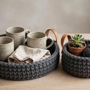 May include: Two dark grey crocheted baskets with tan leather handles. The larger basket holds three speckled ceramic mugs and a folded beige linen napkin. The smaller basket contains a green succulent in a terracotta pot.