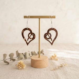 May include: A pair of dangling earrings featuring a dark brown, marbled heart-shaped design with a cutout centre. The earrings are suspended from silver-toned metal hooks and are displayed on a gold-toned metal T-bar stand with a round wooden base.
