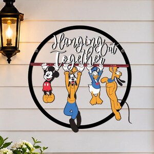 May include: A circular black metal wall sign featuring a red horizontal bar with Mickey Mouse, Goofy, Donald Duck, and Pluto hanging from it. The text "Hanging Out Together" is printed in black script above the characters on the white wall.