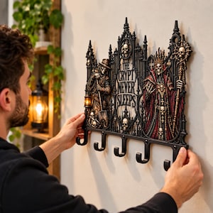 May include: A black wall-mounted key rack featuring a gothic architectural design with intricate skull motifs, a figure holding a shovel, a robed skeletal king holding a staff, and the text THE CRYPT NEVER SLEEPS, complete with five metal hooks.