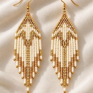May include: A pair of long, dangling earrings featuring a geometric pattern made of small seed beads in shades of cream, tan, and metallic gold. The earrings are suspended from gold-toned metal hooks and have a pointed, fringe-like bottom edge.