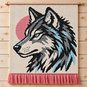 May include: A wall hanging featuring a pixelated wolf head profile in shades of black, gray, and light blue, set against a cream background with a pink circular sun. The piece is suspended from a wooden rod and finished with a pink fringe.