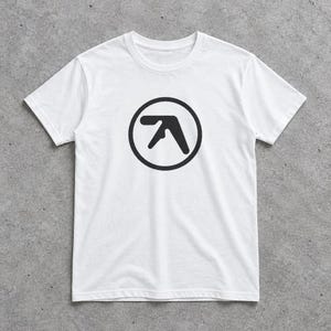 Puede incluir: Una camiseta blanca de manga corta y cuello redondo con un estampado gráfico negro en el pecho. El diseño consiste en una letra A negra estilizada encerrada dentro de un círculo negro sólido, centrado en la parte frontal de la prenda.