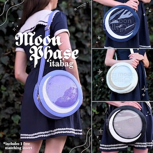 May include: A purple and white round purse with a crescent moon design and a clear plastic window. The purse is worn as a crossbody bag. The text "Moon Phase 'itabag" is visible on the image.