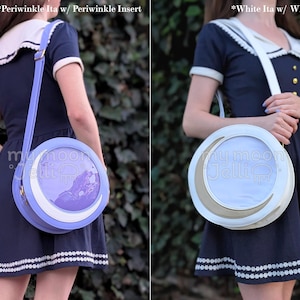 May include: Two round crossbody bags with a crescent moon design. The bag on the left is periwinkle blue with a periwinkle blue insert. The bag on the right is white with a white insert. Both bags have a white strap.