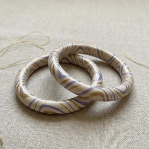 Puede incluir: Dos brazaletes circulares con un acabado liso y brillante con un patrón jaspeado de remolinos en color crema, morado claro y dorado metálico. Las pulseras tienen un perfil redondeado y grueso y están superpuestas.