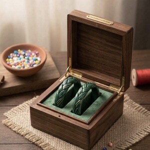 May include: A dark brown wooden box with a hinged lid and a gold-toned metal plaque reading Handcrafted Leatherwear. Inside, two braided dark green leather bracelets rest on a green velvet insert. The box features visible wood grain.