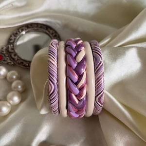 Puede incluir: Un conjunto de cinco brazaletes que incluye una pulsera central trenzada en tonos morado y crema, flanqueada por dos brazaletes lisos en color crema y dos brazaletes exteriores con un patrón de remolinos morados y blancos.