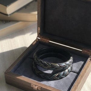 May include: Two dark, braided-style bangles with subtle iridescent highlights resting inside a square, hinged wooden box. The box features a dark gray velvet-lined interior and a metallic latch, providing a secure display for the bracelets.