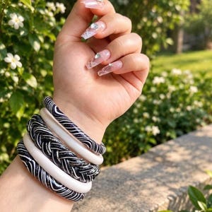 May include: A stack of five bangles worn on the wrist. The set includes two smooth white bangles, two black bangles with thin white swirling lines, and one central black braided bangle with a metallic silver-toned finish.
