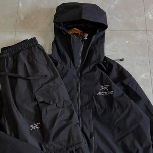 Peut inclure: Un ensemble composé d'une veste à capuche noire et d'un pantalon cargo assorti. La veste présente une fermeture éclair intégrale, une capuche et le logo blanc Arc'teryx sur la poitrine. Le pantalon a une taille élastique avec cordon.