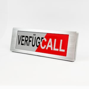 Könnte beinhalten: Ein rechteckiges Schild mit einem Rahmen aus gebürstetem Metall und einem geteilten Hintergrunddesign. Die linke Seite ist hellgrau mit dem schwarzen Text VERFÜG, die rechte Seite ist leuchtend rot mit dem weißen Text CALL, was einen Kontrast bildet.