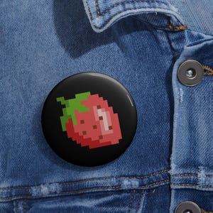 May include: A circular pin-back button featuring a pixel art strawberry design. The strawberry is rendered in shades of red with a green stem and small brown seeds against a solid black background. The button is attached to blue denim fabric.