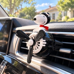 May include: A car air freshener vent clip featuring a white Snoopy figure wearing a black aviator cap and a red scarf, mounted on a black and white propeller-shaped housing that attaches to the horizontal slats of a vehicle dashboard vent.