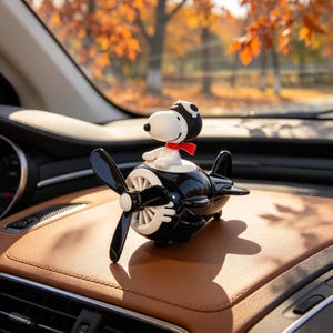 Op de afbeelding: Een wit Snoopy-figuurtje met een zwarte vliegeniersmuts en een rode sjaal, geplaatst op een glanzend zwart speelgoedvliegtuig. Het vliegtuig heeft een opvallende zwarte propeller aan de voorzijde en witte cirkelvormige motordetails aan de zijkanten.