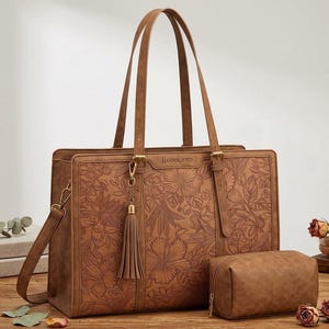 May include: A brown leather tote bag featuring an embossed floral pattern and the word Handcrafted on the front. The bag includes two long shoulder straps, a detachable tassel charm, and a matching rectangular zippered pouch.