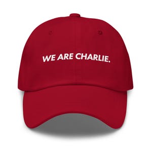 Puede incluir: Gorra de béisbol roja con el texto blanco "WE ARE CHARLIE." La gorra tiene una visera curva y una corona redondeada. El sombrero está hecho de un material suave.