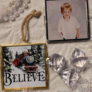 May include: Two square metal-framed hanging ornaments with jute twine loops. One ornament displays a photograph of a young child in a white shirt, while the other features a red train illustration in a snowy forest with the text BELIEVE.
