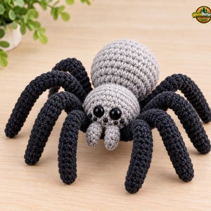 May include: A crocheted spider toy featuring a large, round, light gray abdomen and a smaller light gray head. The spider has eight long, dark charcoal-colored legs and two small gray pedipalps. It features eight black bead eyes arranged in rows.