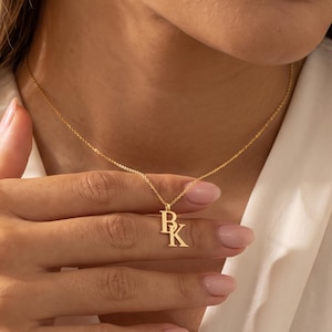 May include: A gold-toned necklace featuring two stacked letter pendants, B and K, suspended from a delicate chain. The letters are rendered in a classic serif font, creating a layered look that hangs centrally against the collarbone area.