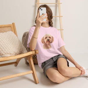 Op de afbeelding: Een lichtroze T-shirt met korte mouwen en een centrale grafische illustratie van een pluizige, lichtbruine hond met hangende oren. Het shirt wordt gedragen met een donkergrijze spijkerbroek. Het ontwerp toont de kop en borst van de hond.