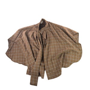May include: A brown cape featuring a white grid pattern throughout. The garment has a high collar, a single button closure at the neck, and a wide, flowing silhouette that drapes outward from the shoulders, creating a structured yet loose fit.