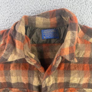 Op de afbeelding: Een overhemd met lange mouwen en knoopsluiting met een ruitpatroon in tinten oranje, bruin en beige. Het overhemd heeft een puntige kraag en een donkerbruine voering. Een blauw Pendleton-label met witte tekst is in de nek genaaid.