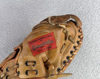 Könnte beinhalten: Ein beigefarbener Baseballhandschuh aus Leder mit einem rechteckigen roten Aufnäher, der den Text "DESIGNED IN UNITED STATES BY Johnny Walker MADE IN JAPAN" trägt, akzentuiert durch schwarze Metallösen und Lederbänder.