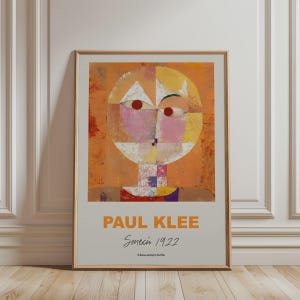 May include: A framed art print featuring Paul Klee's Senecio 1922 painting. The artwork displays a stylised, geometric human face in shades of orange, yellow, and pink with two red circular eyes. Below the image, the text reads PAUL KLEE Senecio 1922.