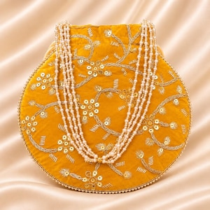 May include: A round mustard yellow velvet handbag featuring intricate gold thread embroidery and sequin floral patterns. The bag is adorned with a multi-strand white pearl beaded handle and a delicate pearl beaded trim along the circular edge.