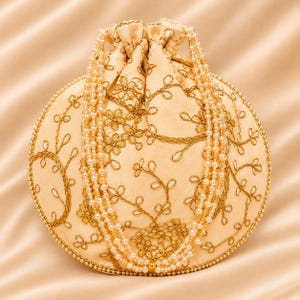 May include: A round, cream-colored drawstring pouch featuring intricate gold floral embroidery and a beaded trim. The bag is adorned with a long, multi-strand pearl necklace draped over the front, adding a layered texture to the design.