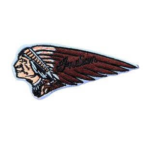 May include: An embroidered patch featuring a profile of a person wearing a traditional headdress with long feathers extending to the right. The design includes the word Indian in black script lettering across the brown feathers and a light blue border.