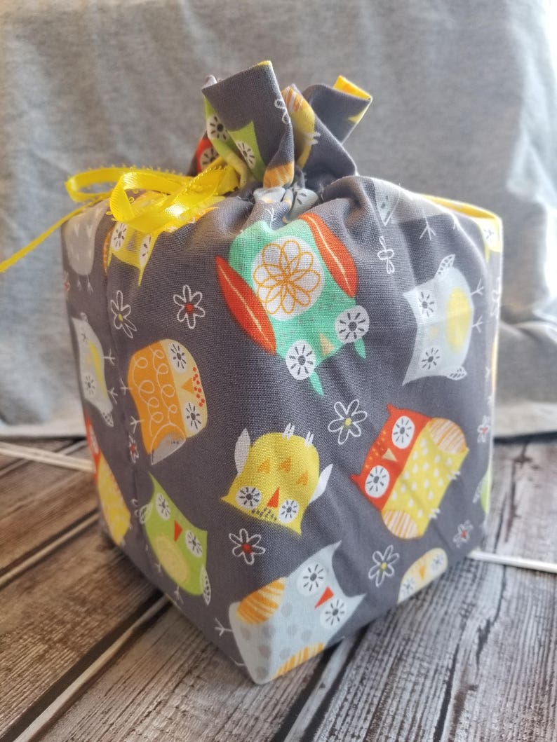 Puede incluir: Una caja de almacenamiento de tela gris con una cinta amarilla atada alrededor. La caja est&aacute; decorada con un patr&oacute;n de b&uacute;hos coloridos y flores blancas.