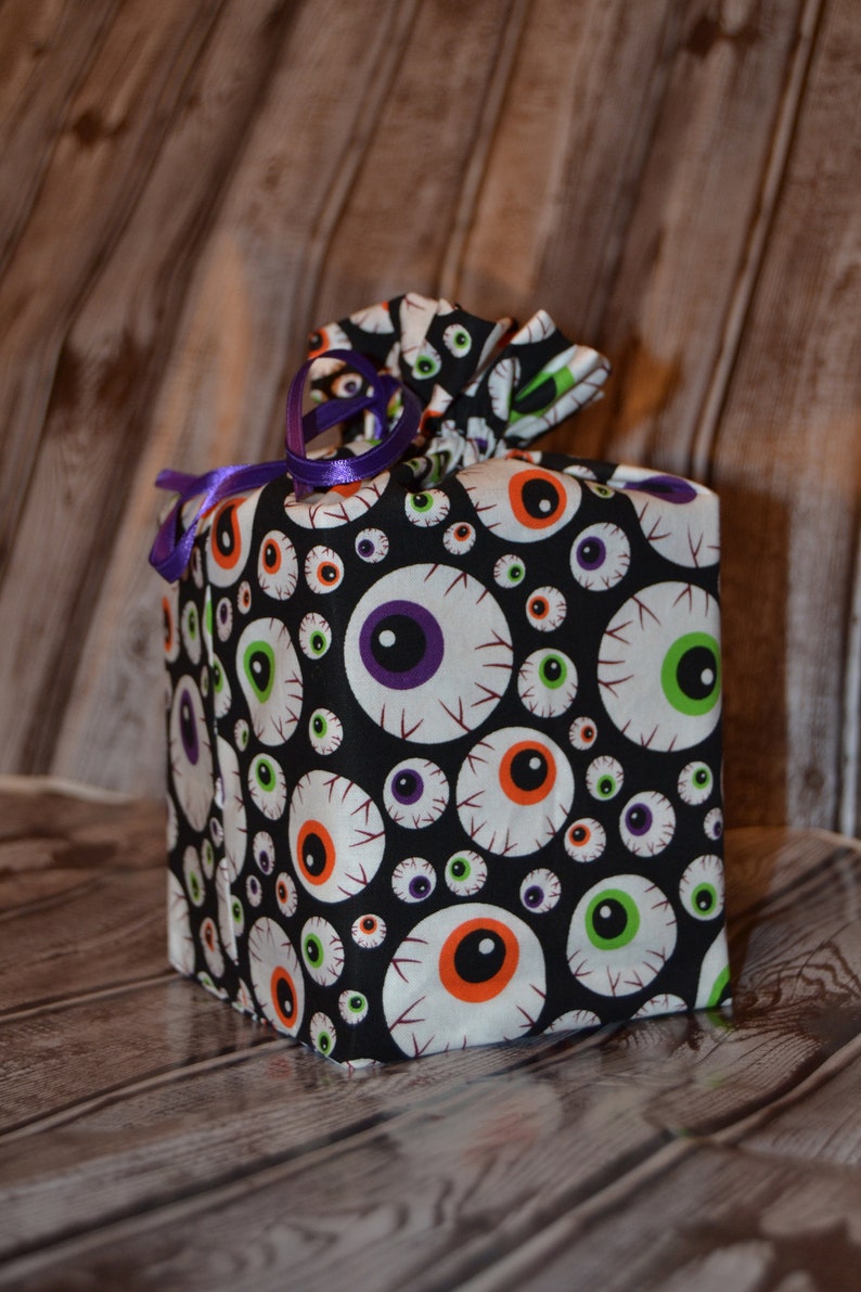 May include: A black fabric gift bag with a white and colorful eyeball pattern. The bag is tied with a purple ribbon.