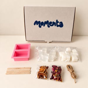 Può includere: Un kit per il fai da te in una scatola di cartone con la scritta "moments" contenente uno stampo in silicone rosa, due sacchetti di cubetti di sapone trasparenti e bianchi, quattro bastoncini di legno, bustine di arance essiccate e petali.
