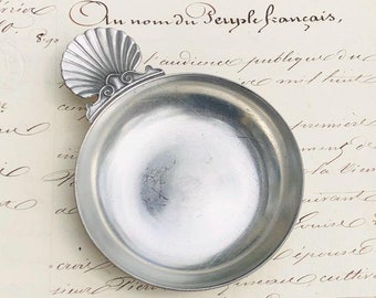 Peut inclure: Un plat de service antique en argent, en forme de coquille Saint-Jacques. Le plat a une surface lisse et réfléchissante et repose sur un morceau de papier avec du texte manuscrit en français.