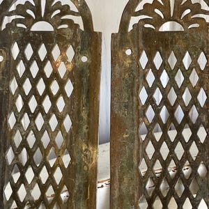 May include: Two antique, weathered metal panels with a diamond lattice pattern and ornate, arched tops. The tops feature a stylized sunburst design. The panels have a distressed, aged appearance with hints of green and brown.