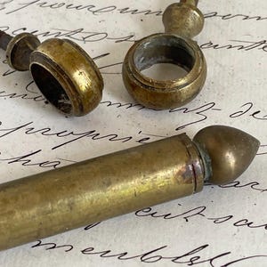 Op de afbeelding: Twee messing ringgrepen met schroefdraad en een cilindrische messing staaf met een puntige eindknop. De metalen onderdelen vertonen een verweerde, verouderde patina met zichtbare tekenen van oxidatie en slijtage aan het oppervlak.