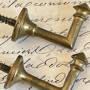 Può includere: Due ganci antichi a forma di L in ottone con viti. I ganci hanno una parte superiore decorativa a punta e una base circolare. L'ottone ha un aspetto leggermente invecchiato e ossidato. Lo sfondo è una carta vintage con scritte a mano.