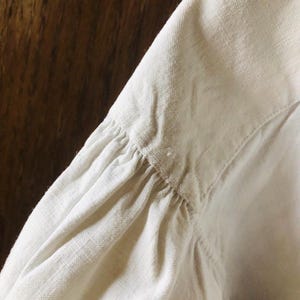 May include: Close-up of the white linen fabric of a shirt sleeve, showing the gathered detail at the shoulder seam.