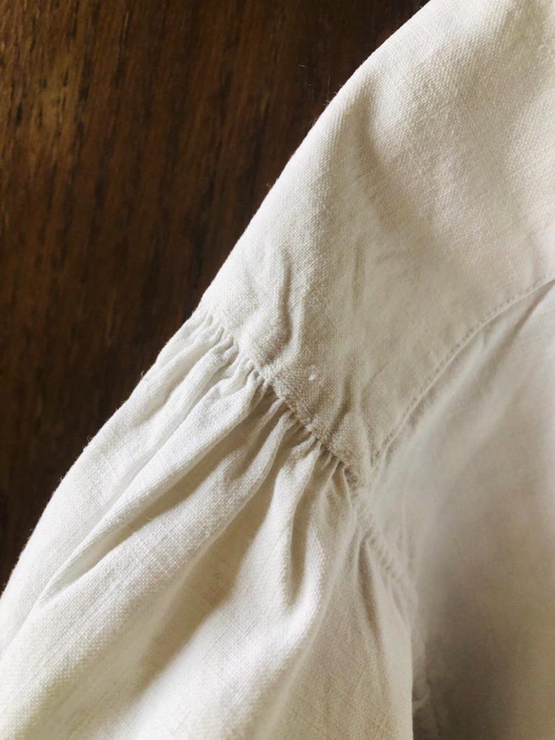 May include: Close-up of the white linen fabric of a shirt sleeve, showing the gathered detail at the shoulder seam.