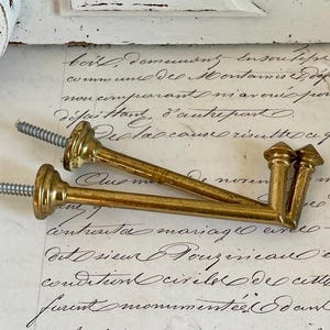 Può includere: Due supporti per bastone per tende in ottone vintage con viti. I supporti hanno un design ornato e sono di colore oro caldo. Le viti sono argentate. I supporti poggiano su un pezzo di carta antica con scritte a mano.