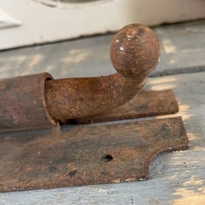 May include: Close-up of a rusty, antique metal latch. The latch features a rounded ball handle and a rectangular base with a single hole. The metal has a weathered, aged appearance, with visible rust and discoloration.