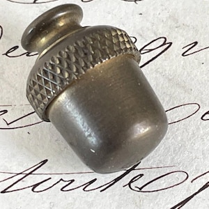 May include: A small, antique-style metal acorn-shaped object. The acorn has a textured cap and a smooth, rounded body. The metal has a tarnished, aged appearance, suggesting it is vintage or vintage-inspired. The acorn is a decorative item.