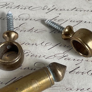 Puede incluir: Primer plano de tres herrajes de muebles antiguos de latón. Cada pieza presenta un diseño redondo en forma de anillo con un tornillo adjunto. El latón tiene una pátina cálida y envejecida, lo que sugiere orígenes vintage. El fondo es un papel vintage con escritura manuscrita.